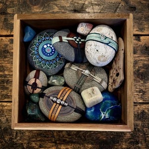 May include: A square wooden box containing a collection of smooth river stones, some wrapped in intricate wire and leather cord designs, others painted with blue and green mandala patterns, alongside small polished gemstones and a piece of driftwood.
