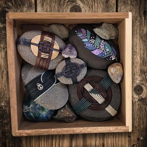 May include: A square wooden box containing several smooth gray stones. Some stones are wrapped in woven leather and cord in brown, black, and cream colors. One stone features a painted purple and teal feather design, and another has a small silver peace sign charm attached.