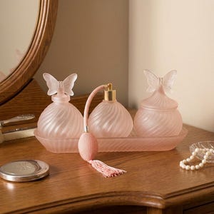 May include: A set of three frosted pink glass containers with spiral patterns and butterfly-shaped stoppers, arranged on a matching rectangular tray. One container features a pink fabric atomizer bulb with a tassel, alongside a hairbrush and pearls.