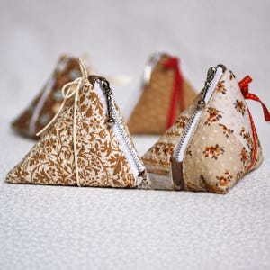 May include: Three triangular fabric pouches with zippers. The front pouch features a brown floral pattern on a cream background with a white cord tie. The right pouch has a beige floral print and a red ribbon. The back pouch is solid brown.