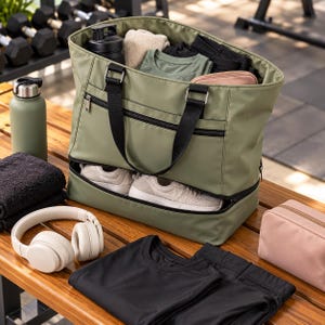 Pode incluir: Uma bolsa de desporto verde-oliva com alças pretas e um compartimento inferior separado contendo um par de ténis claros. A bolsa está cheia de roupas, uma garrafa de água preta e uma pequena bolsa, com auscultadores e roupas pretas por perto.