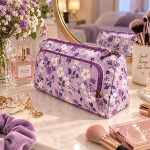 Puede incluir: Un neceser rectangular de tela color violeta claro con un patrón repetitivo de flores blancas y violeta oscuro con tallos finos. El estuche cuenta con un compartimento principal con cremallera y un bolsillo frontal más pequeño con herrajes bronce.