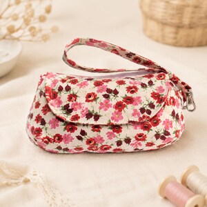Könnte beinhalten: Eine kleine Handgelenktasche mit cremefarbener Basis und einem lebendigen Blumenmuster aus roten, rosafarbenen und dunkelburgunderroten Blüten. Die Tasche hat einen abgerundeten Klappenverschluss und eine passende Handschlaufe mit silbernem Metallverschluss.