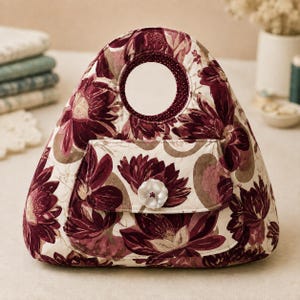 May include: A triangular quilted handbag featuring a dark burgundy and cream floral pattern. The bag has a circular cutout handle and a front flap pocket secured with a single white flower-shaped button. The fabric has a soft, padded texture.