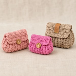 May include: Three small crochet bags in light pink, bright pink, and beige. Each bag features a textured stitch pattern, a fold-over flap, and a single wooden button closure. The beige bag includes a small brown leather-like tab on the flap.