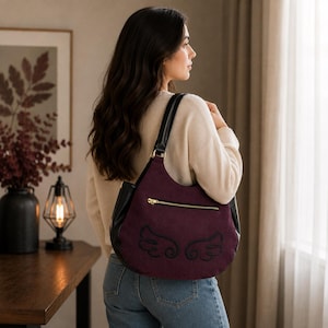 Peut inclure: Un sac à bandoulière de style hobo avec un corps en suédine bordeaux profond, des panneaux latéraux et des bretelles en similicuir noir. Le sac présente une poche zippée dorée horizontale et un motif d'ailes brodé en noir sur l'extérieur.