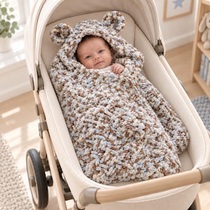 May include: A chunky crochet baby swaddle blanket featuring a hood with two small rounded ears. The yarn is a variegated blend of light blue, cream, and tan colors. The swaddle is wrapped around an infant, covering the body while leaving the face visible.