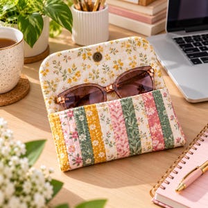 Peut inclure: Un étui à lunettes en tissu matelassé avec un motif à rayures verticales de fleurs dans des tons de jaune, vert et rose. L'étui possède un rabat repliable avec une fermeture à pression en métal et contient une paire de lunettes de soleil teintées.