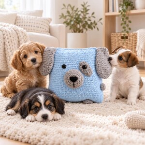 May include: A square, light blue crocheted pillow shaped like a dog, featuring gray crocheted ears, a gray snout, a black nose, and black button eyes. The pillow is surrounded by three small puppies: one golden-brown, one black, white, and brown, and one white and brown.
