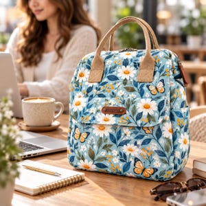 Può includere: Uno zaino azzurro con un motivo di margherite bianche, foglie verdi e farfalle arancioni. La borsa presenta due manici superiori in tessuto beige, una tasca frontale con una toppa rettangolare marrone e una chiusura a zip con linguetta in pelle.