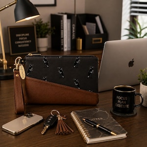 Könnte beinhalten: Eine rechteckige Handgelenktasche mit einem schwarzen Stoffoberteil mit wiederkehrendem weißem Rabenmuster und einem unteren Teil aus einfarbigem braunem Kunstleder. Die Tasche verfügt über eine passende braune Handschlaufe mit goldenem Verschluss.