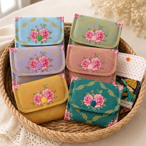 May include: A collection of small fabric wallets featuring a floral pattern with pink roses and green leaves. Each wallet has a flap closure secured with a colourful plastic snap button. The wallets are available in blue, green, lavender, pink, yellow, and teal.