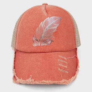 May include: A coral-coloured baseball cap with a distressed, frayed brim and a mesh back. The front panel features a metallic silver feather graphic. The cap has a washed-out, vintage appearance with visible stitching and a structured crown design.