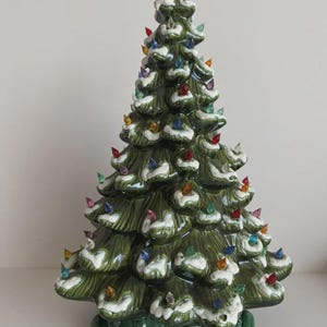 May include: A glossy green ceramic Christmas tree figurine featuring white snow-like accents on the branches and numerous small, multi-colored translucent plastic light bulbs inserted throughout the structure, resting on a decorative green base.