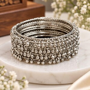 May include: A set of silver-toned metal bangles featuring a stacked design. The outer bangles are adorned with numerous small, dangling spherical charms that create a textured, layered appearance. The inner bangles have a smooth, polished finish.