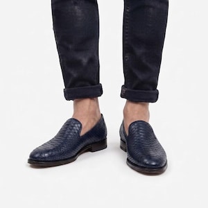 May include: A pair of dark blue loafers with a textured snakeskin-like pattern on the upper, featuring a low-cut silhouette and a small stacked heel, worn with dark blue cuffed denim jeans that end just above the ankle.