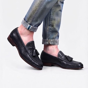 May include: A pair of black loafers with a textured snakeskin pattern and a tassel detail on the top. The shoes feature a low heel and a dark brown sole, worn with light blue distressed denim jeans that are cuffed at the bottom hem.
