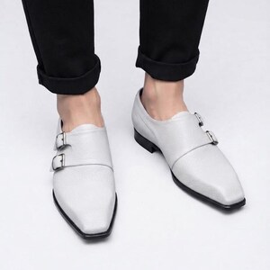 A top-down view of a person's lower legs wearing black cuffed trousers and a pair of white pebble-grain leather double monk strap shoes, standing on a grey floor.