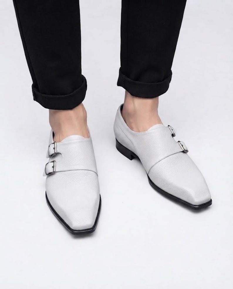 A top-down view of a person's lower legs wearing black cuffed trousers and a pair of white pebble-grain leather double monk strap shoes, standing on a grey floor.