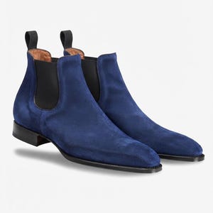 Pode incluir: Um par de botas Chelsea de camurça azul com painéis laterais elásticos pretos, puxadores pretos no calcanhar e um salto baixo preto com sola preta lisa. As botas têm um design de bico fino e um forro interior em couro castanho claro.