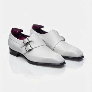 A pair of the same white pebble-grain leather double monk strap shoes presented at a three-quarter angle on a reflective white studio floor, with distinct purple-stained wooden shoe trees inserted