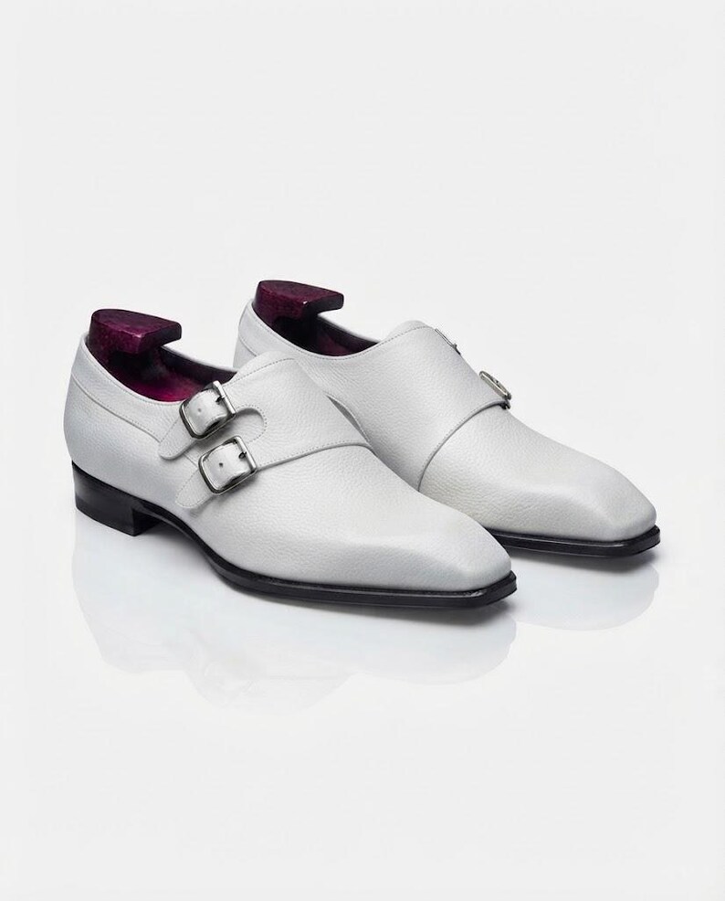 A pair of the same white pebble-grain leather double monk strap shoes presented at a three-quarter angle on a reflective white studio floor, with distinct purple-stained wooden shoe trees inserted
