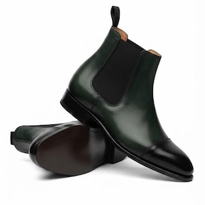 May include: A pair of dark forest green leather Chelsea boots featuring a polished black cap toe, black elastic side panels, a black pull tab at the heel, and a dark brown sole. The boots have a smooth finish and a structured ankle-high silhouette.