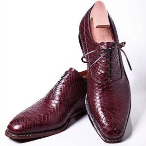 Könnte beinhalten: Ein Paar dunkelburgunderfarbene Oxford-Schuhe mit strukturiertem Schlangenledermuster, spitzer Zehenpartie mit dekorativen Brogue-Perforationen, dünnen dunklen Schnürsenkeln und einem Holzschuhspanner im aufrechten Schuh.