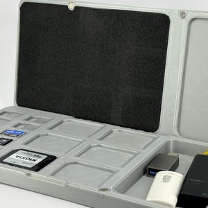 Puede incluir: Un estuche de almacenamiento de plástico gris claro con tapa abatible y forro interior de espuma negra. La base contiene múltiples ranuras rectangulares para tarjetas de memoria, incluida una con la etiqueta KIOXIA, y un compartimento.