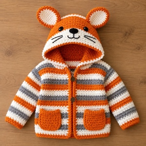 May include: A crocheted hooded jacket featuring a fox face design on the hood with orange, white, and gray horizontal stripes. The garment includes two front pockets, a zipper closure with two brown buttons, and rounded ears on top of the hood.