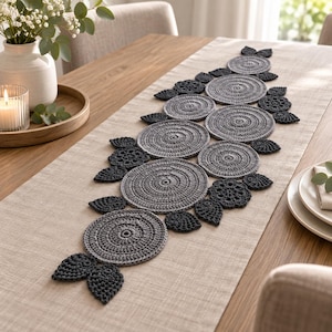 May include: A beige linen table runner featuring a central decorative arrangement of crocheted elements. The design includes multiple light gray circular motifs and dark gray leaf-shaped accents, all stitched together in a linear pattern.