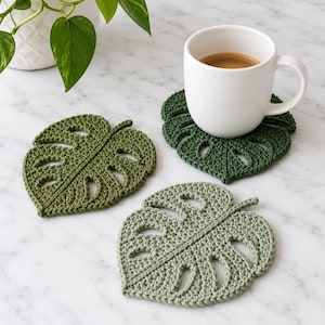 Peut inclure: Trois dessous de verre au crochet en forme de feuilles de monstera dans des nuances de vert variées. Deux dessous de verre sont vert foncé et un est vert sauge plus clair. Un dessous de verre vert foncé soutient une tasse en céramique blanche.
