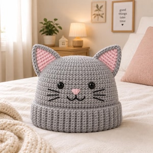 May include: A gray crocheted beanie featuring a cat face design with pointed ears, pink inner ear details, black button eyes, a small pink nose, and embroidered black whiskers. The hat has a thick, ribbed brim and a textured, solid gray crown.