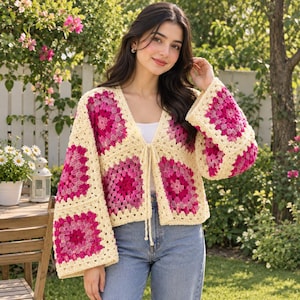May include: A crochet cardigan featuring a granny square pattern in shades of cream, magenta, and light pink. The garment has long, wide sleeves, a V-neckline, and a front tie closure made of matching cream yarn, worn over a white top and blue jeans.