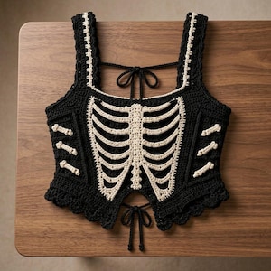 Op de afbeelding: Een zwarte gehaakte crop top met een crèmekleurig ribbenkastontwerp aan de voorzijde en kleine botmotieven aan de zijkanten. Het kledingstuk heeft brede schouderbanden, een geschulpte zoom en verstelbare striksluitingen aan de rug en onderzijde.