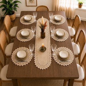 May include: A rectangular wooden dining table set for six with cream-colored crocheted placemats and a matching oval table runner. Each place setting includes a white bowl on a plate, a clear glass, and a fork. A vase with dried flowers sits centrally.