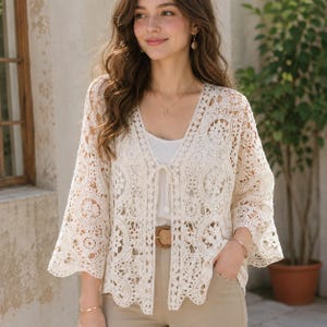 May include: A cream-coloured, open-front crochet cardigan featuring intricate circular floral patterns, three-quarter length bell sleeves, and a delicate tie closure at the centre neckline. The garment is worn over a plain white top and tan trousers.