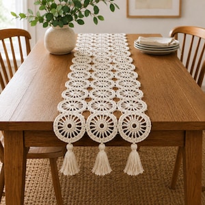 May include: A long, off-white crochet table runner featuring a repeating circular pattern with intricate spokes. The runner is finished with three decorative tassels hanging from the bottom edge, creating a textured and detailed textile design.