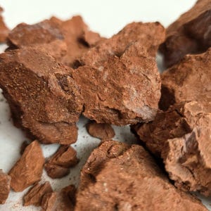 Puede incluir: Una colección de rocas de arcilla crudas y dentadas en un tono marrón rojizo intenso. Las piedras presentan una textura rugosa, porosa e irregular con depósitos minerales visibles y finas partículas de polvo sobre los fragmentos.