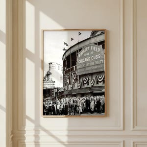 Puede incluir: Una impresión enmarcada en blanco y negro de Wrigley Field, hogar de los Chicago Cubs. La imagen muestra una multitud reunida fuera del estadio con un gran letrero que dice Wrigley Field Home of Chicago Cubs Games Start One Thirty National League Champions.