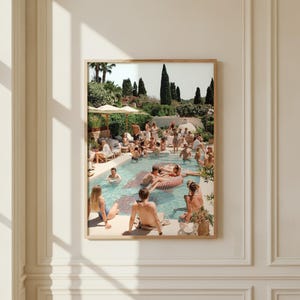 Könnte beinhalten: Ein gerahmter Wandkunstdruck, der eine sonnendurchflutete Außenpoolszene zeigt, in der sich Menschen im und am Wasser entspannen. Das Bild zeigt beige Sonnenschirme, üppige grüne Bäume und einen Steinterrassenbereich unter einem hellen, klaren Himmel.