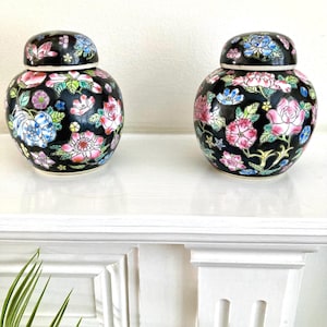 May include: A pair of round ceramic ginger jars with lids, featuring a glossy black background decorated with a vibrant floral pattern in shades of pink, blue, green, and yellow. Each jar has a rounded body and a matching removable lid.