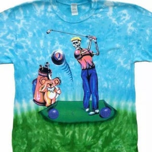 May include: A tie-dye t-shirt with a blue and green color scheme. The shirt features a cartoon skeleton golfer with a bear in a golf bag. The bear is wearing a pink shirt and a brown hat. The skeleton is wearing a red shirt, blue pants, and a yellow hat. The shirt has a tie-dye pattern in shades of blue and green.