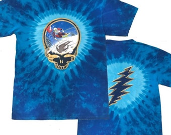 Może przedstawiać: Niebieska koszulka tie-dye z grafiką czaszki Steal Your Face zespołu Grateful Dead, przedstawiającą narciarza w zimowej scenerii. Na plecach koszulki znajduje się logo błyskawicy zespołu Grateful Dead.