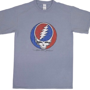 May include: A light blue t-shirt with a vintage-style Grateful Dead Steal Your Face logo in red, white, and blue. The logo is a skull with a lightning bolt in a circle.