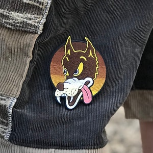 May include: Brown corduroy shorts with a patch of a cartoon wolf with a yellow background. The shorts have a beige trim and are made of a patchwork fabric.