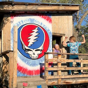 May include: A wooden treehouse with a red, white, and blue tie-dye tapestry featuring a Grateful Dead Steal Your Face logo. Two children are standing on the platform of the treehouse, smiling and looking at the camera.