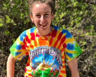 May include: A woman with dreadlocks is wearing a tie-dye t-shirt with the Grateful Dead logo. The shirt is a mix of red, orange, yellow, blue, and green.