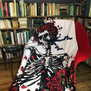 May include: White tapestry featuring a skeleton with a skull wearing a crown of red roses. The skeleton is surrounded by red roses and green leaves. The tapestry is draped over a red chair.