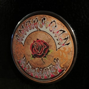 May include: A round button with a gold and brown background featuring a red rose with green leaves. The words "The Grateful Dead" are written in a stylized font around the rose.